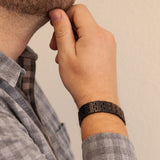 Male model wearing the Brilliant Black matte stainless steel bracelet, hand resting on chin to display engraved matte links and modern masculine style.
