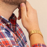 Male model wearing the Golden Light matte gold stainless steel bracelet, hand resting near chin to display elegant gold checkered design.